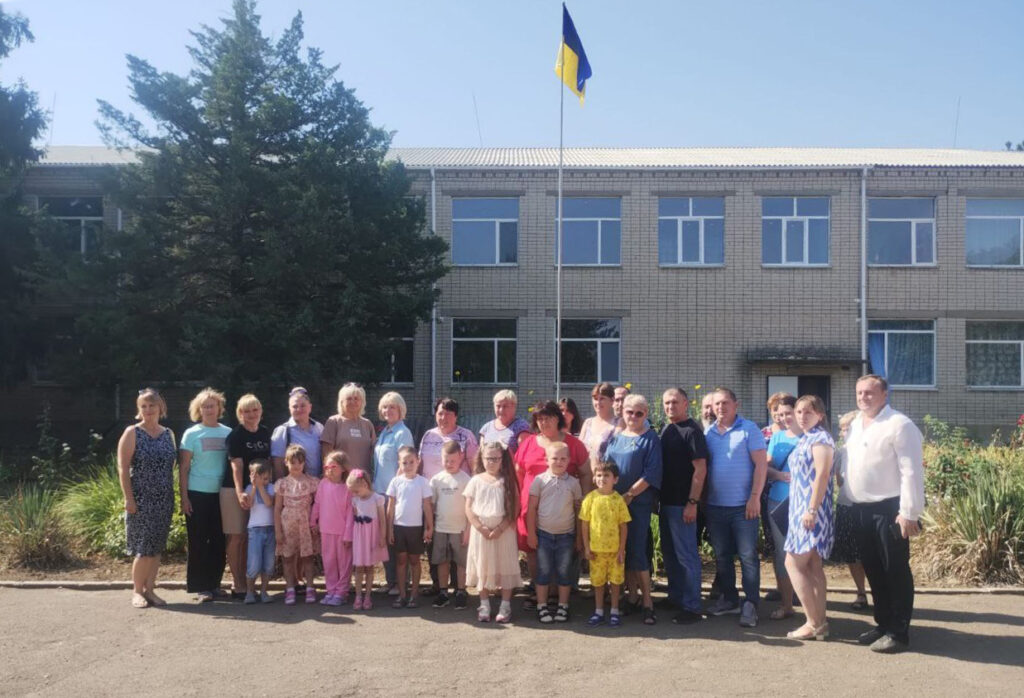 Група дорослих і дітей стоїть перед цегляною будівлею з кількома вікнами. На флагштоку — прапор України. Люди вишикувались у ряд, позують для фотографії. Навколо — дерева та зелень, схоже на територію школи або громадської установи.