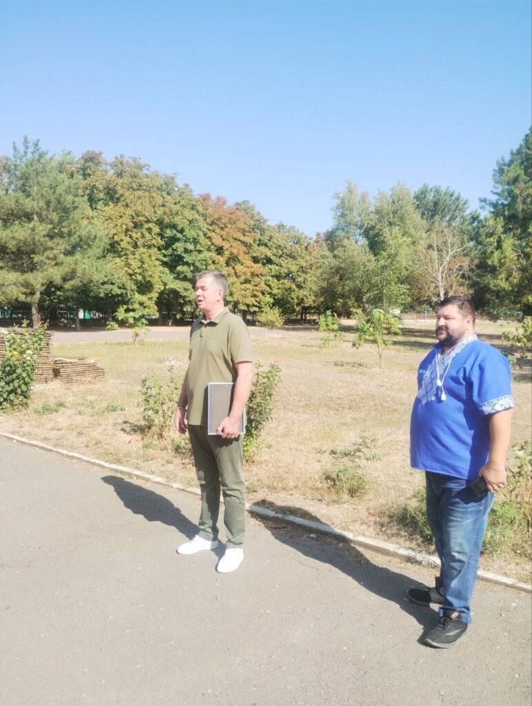 Двоє людей стоять на мощеній ділянці поруч із зеленою зоною. Один тримає ноутбук або записник, інший — смартфон. На задньому плані — дерева з зеленим і жовтогарячим листям, що свідчить про осінню пору.