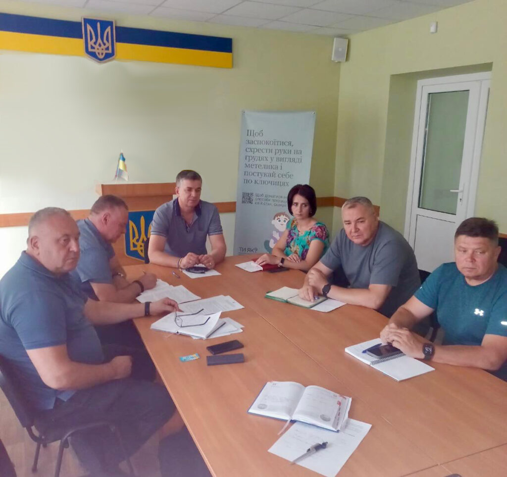 ChatGPT сказал: На фото шість людей сидять за великим столом у світлій кімнаті з українською символікою на стіні. Перед ними розкладені документи, блокноти й телефони. Вони зосереджено обговорюють питання на нараді, деякі роблять записи. Атмосфера офіційна та робоча.