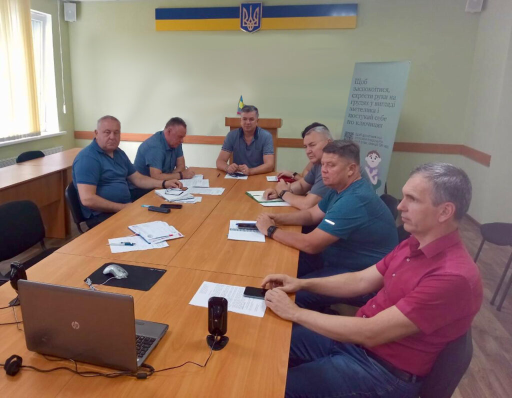 ChatGPT сказал: На фото шість людей сидять за великим столом у світлій кімнаті з українською символікою на стіні. Перед ними розкладені документи, блокноти й телефони. Вони зосереджено обговорюють питання на нараді, деякі роблять записи. Атмосфера офіційна та робоча.