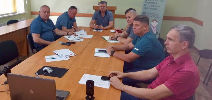 ChatGPT сказал: На фото шість людей сидять за великим столом у світлій кімнаті з українською символікою на стіні. Перед ними розкладені документи, блокноти й телефони. Вони зосереджено обговорюють питання на нараді, деякі роблять записи. Атмосфера офіційна та робоча.