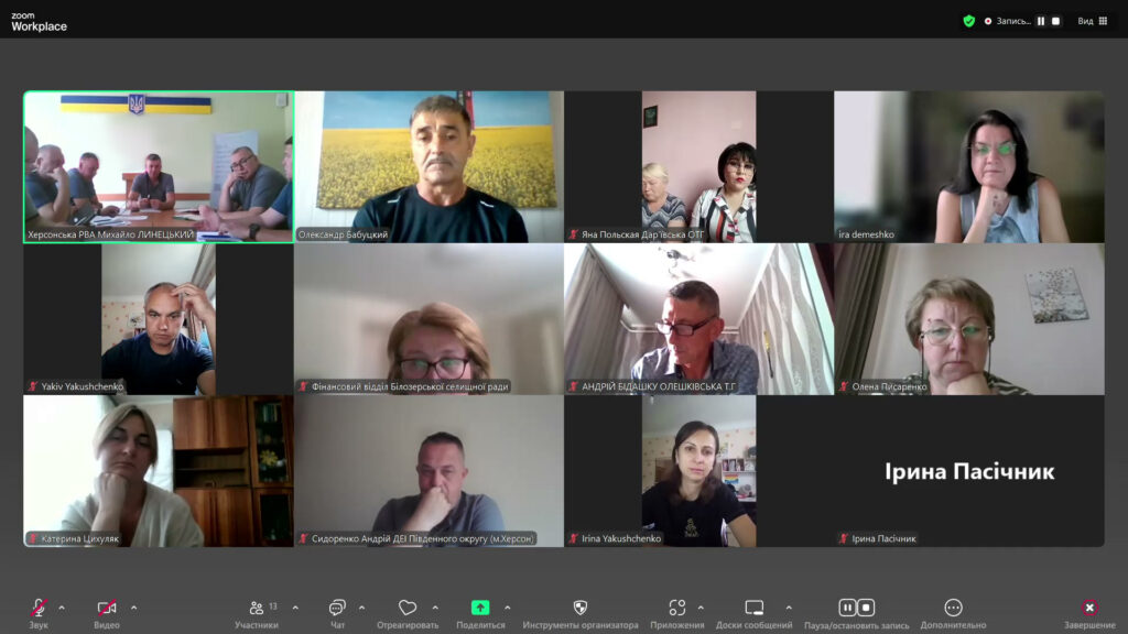 На зображенні відкрите вікно Zoom-конференції. У ній бере участь понад десять людей, розділених на окремі віконця. У верхньому лівому кутку — група чоловіків у кімнаті з українським прапором, серед них начальник Херсонської РДА (ВЦА) Михайло Линецький. Інші учасники підключені з різних місць: хтось сидить вдома, інші в офісах. Більшість виглядають зосередженими, деякі роблять записи. Атмосфера ділова — йде робоча онлайн-нарада.