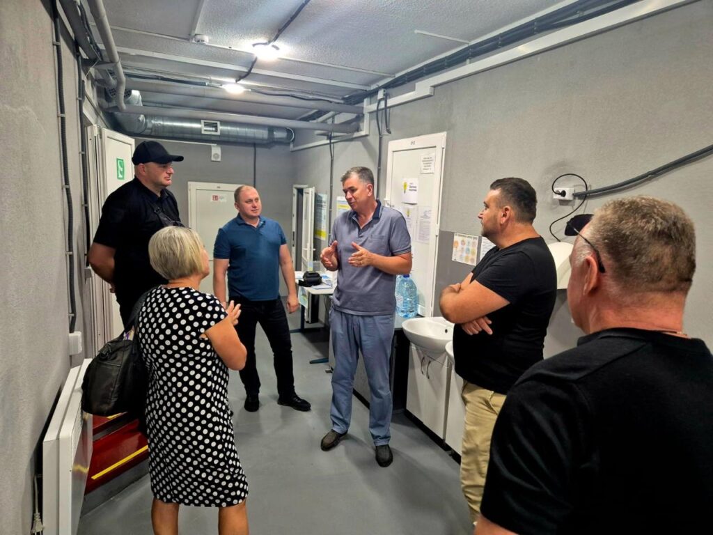 Шестеро людей обговорюють щось у вузькому коридорі з сірими стінами, трубами на стелі та дверима з табличкою «Вихід».