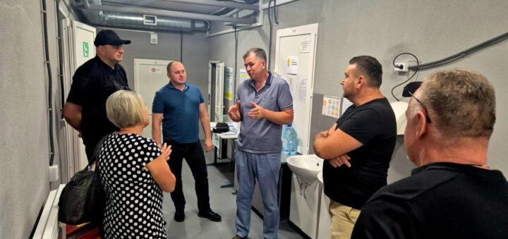 Шестеро людей обговорюють щось у вузькому коридорі з сірими стінами, трубами на стелі та дверима з табличкою «Вихід».