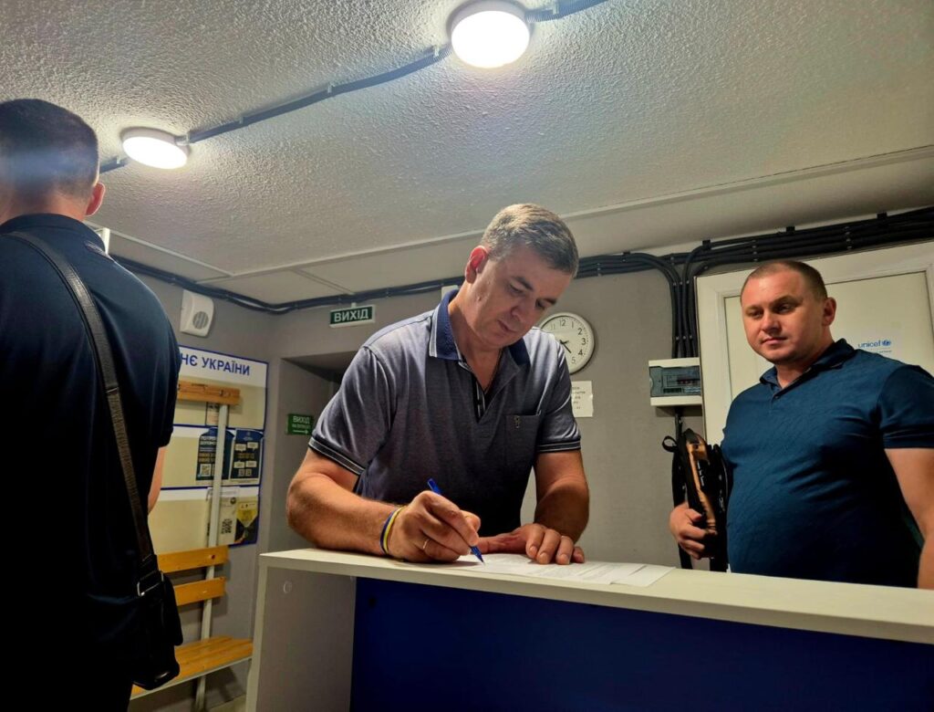 Троє людей біля стійки в адміністративному приміщенні; один пише, інші спостерігають. На стіні — таблички «ВИХІД» і «СЛУЖБА УКРАЇНИ», годинник показує 14:34.