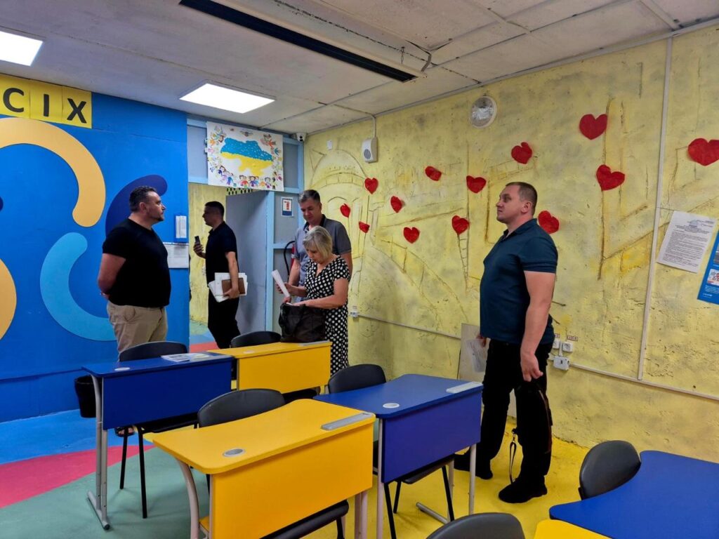 Кольорова шкільна класна кімната з чотирма людьми, які розмовляють; на стінах — карта України, серця та яскраві візерунки.