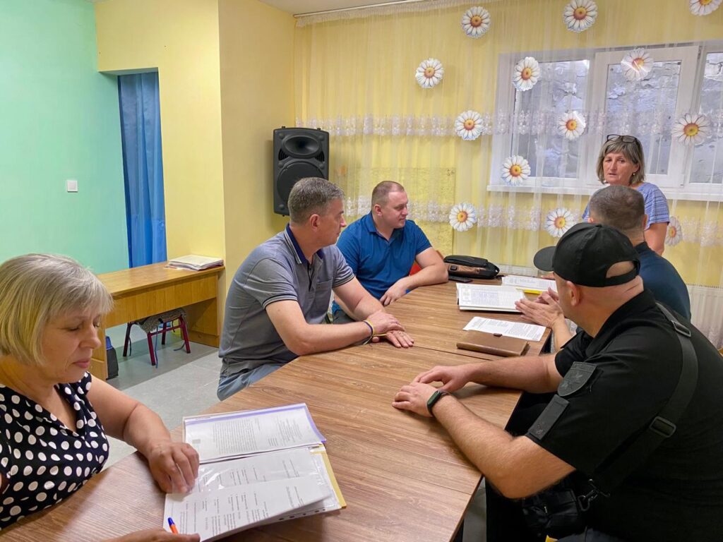 На фото кілька людей сидять за дерев’яним столом у кімнаті з жовтими стінами та шторами з квітковим орнаментом. Вони ведуть робочу розмову, перед ними лежать документи й папки. Одна жінка стоїть і звертається до присутніх, решта уважно слухають