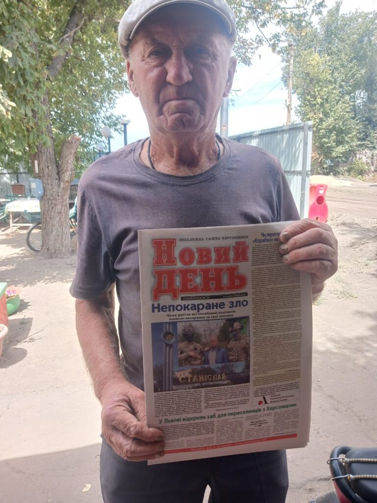 Газета «Новий День» із заголовком «Непокаране зло» лежить у руках людини. На першій сторінці — фото пам’ятника та групи людей з підписом «Станіслав». Нижче — заголовок «У Львові відкрили хаб для переселенців з Херсонщини». На фоні — дерева, паркан і предмети на подвір’ї.