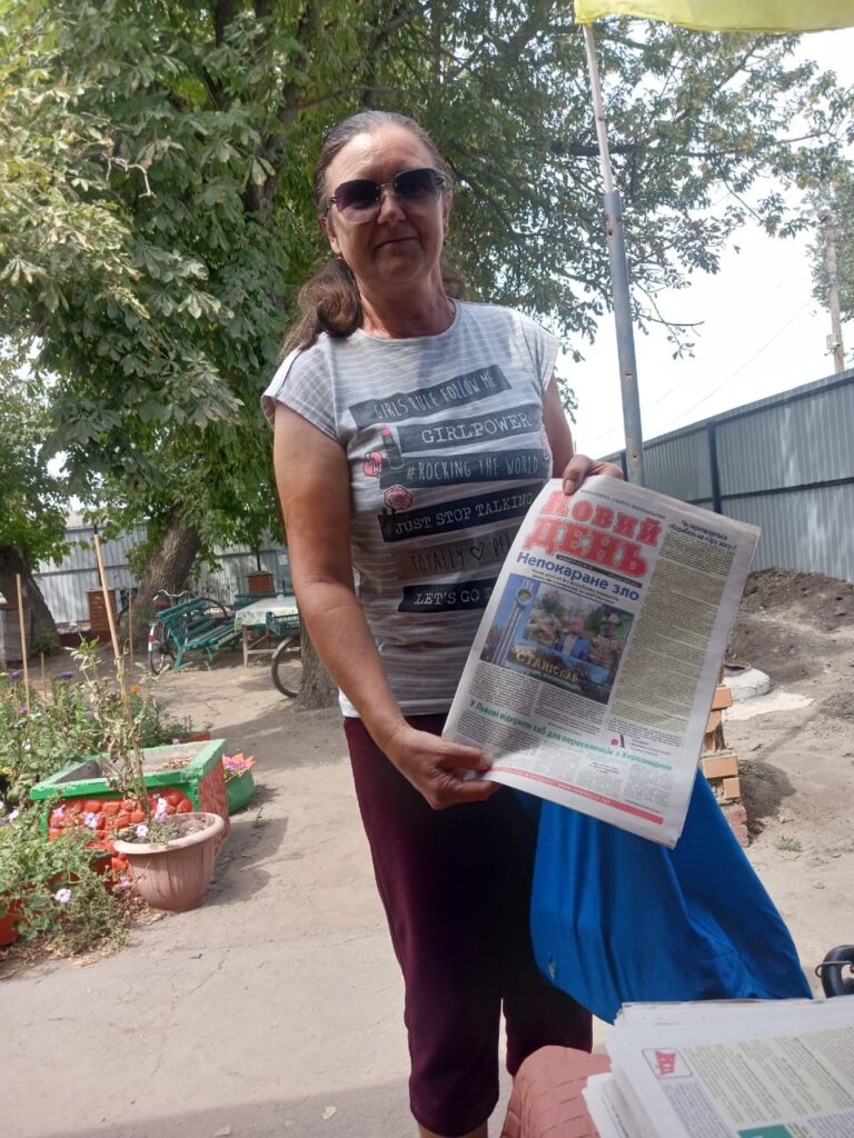 Людина стоїть на подвір’ї, тримаючи газету «Новий день». Вона одягнена у сіру футболку з написами «GIRLPOWER», «#ROCKING THE WORLD» і «JUST STOP TALKING». На фоні — дерево, паркан і горщики з рослинами.