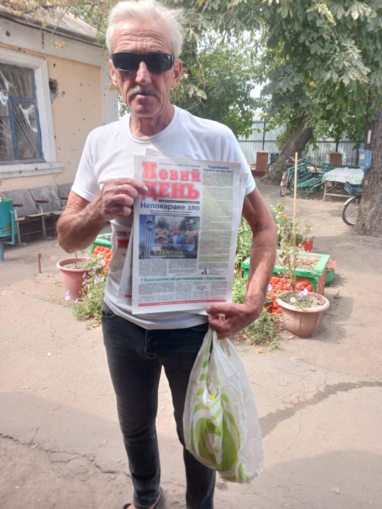 Чоловік стоїть на подвір’ї біля будинку, тримає газету «Новий день» із заголовком «Непокаране зло» та прозорий пакет. Він одягнений у білу футболку й темні штани. На фоні — вікно, рослини в горщиках і дерево.