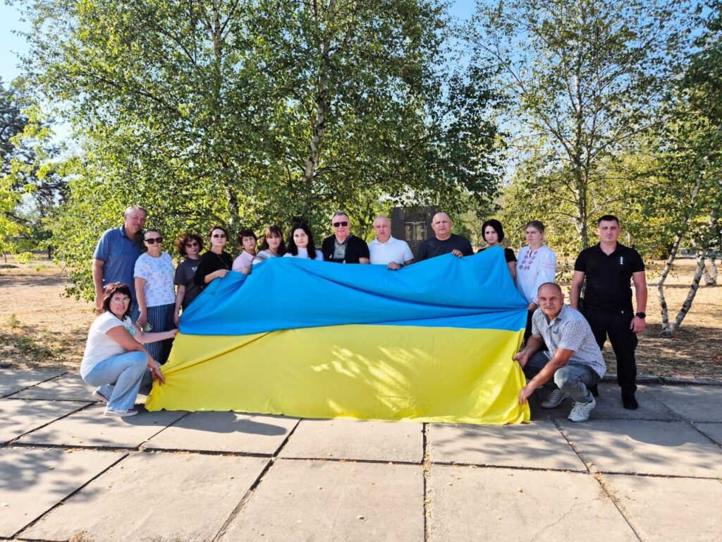 Група людей на вулиці тримає великий прапор України. Дехто стоїть, дехто — на колінах, щоб розгорнути полотно. Навколо — дерева та зелень, схоже на парк або сквер.