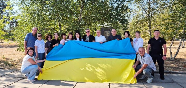 Група людей на вулиці тримає великий прапор України. Дехто стоїть, дехто — на колінах, щоб розгорнути полотно. Навколо — дерева та зелень, схоже на парк або сквер.