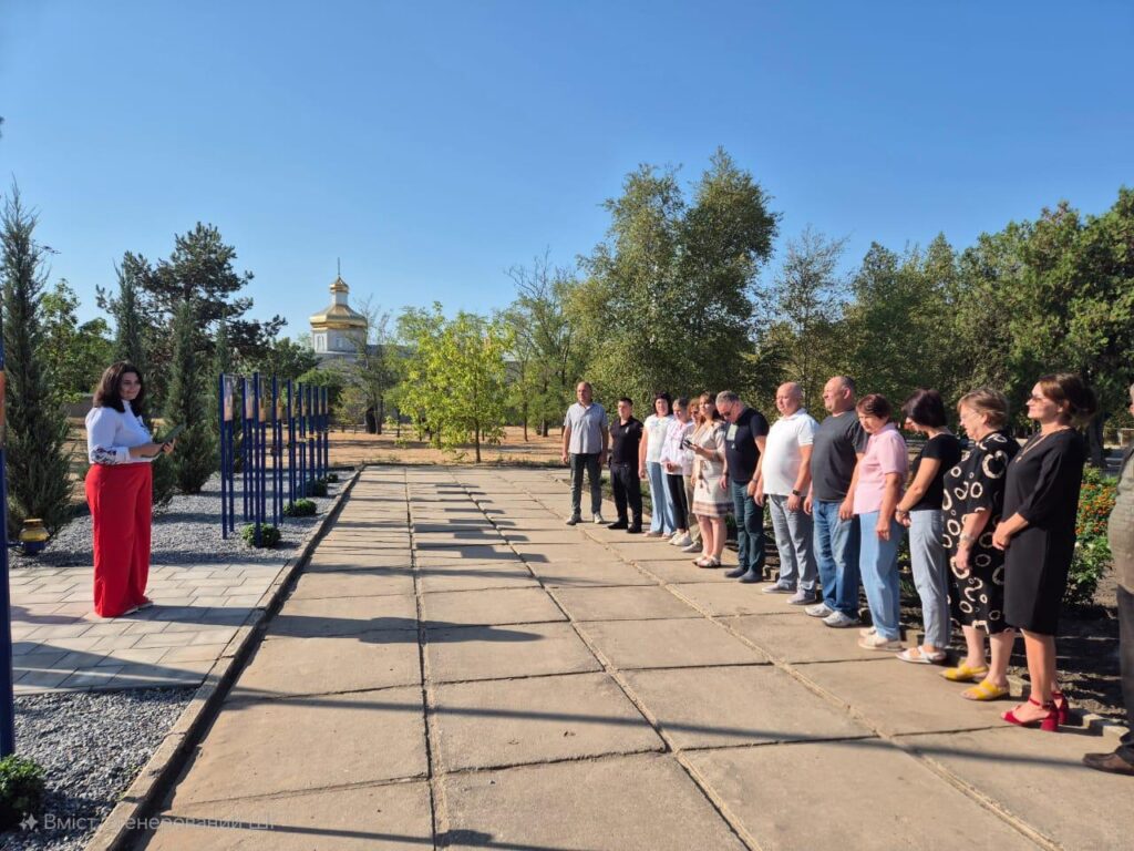 рупа людей стоїть на мощеній доріжці просто неба, слухаючи жінку, яка звертається до них. На задньому плані — церква або каплиця з золотим куполом, а також сині металеві конструкції з табличками. Навколо — дерева, кущі та чисте блакитне небо.