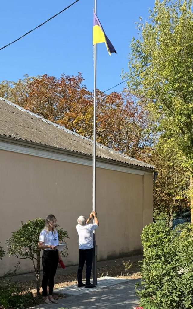 Двоє людей стоять біля флагштока просто неба. Один піднімає жовто-блакитний прапор України, інший тримає папери та червоний предмет. На задньому плані — будівля з хвилястим дахом і дерева з осіннім листям. Сонячна погода.