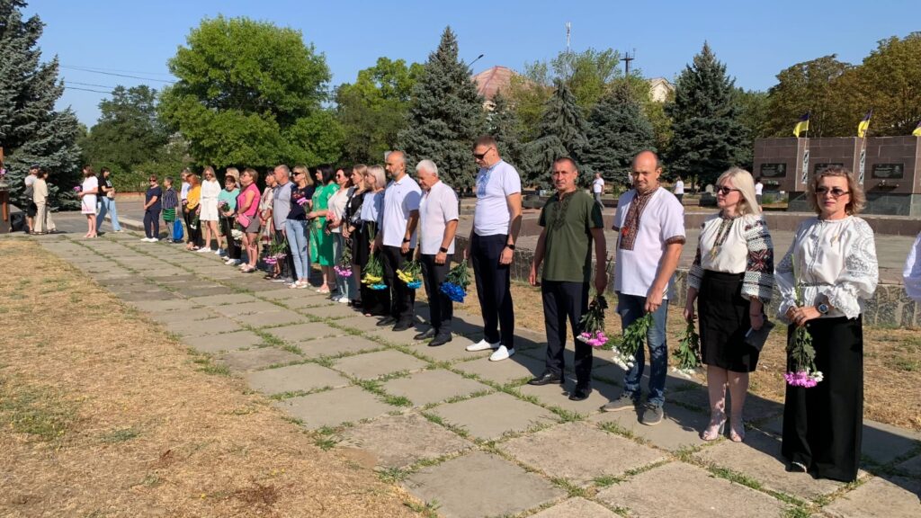 Група людей стоїть у ряд на мощеній доріжці в парку, тримаючи квіти. Позаду — пам’ятники та прапори, що свідчить про меморіальну або урочисту подію. Деякі учасники вдягнені в традиційний одяг. Навколо — зелень і дерева.