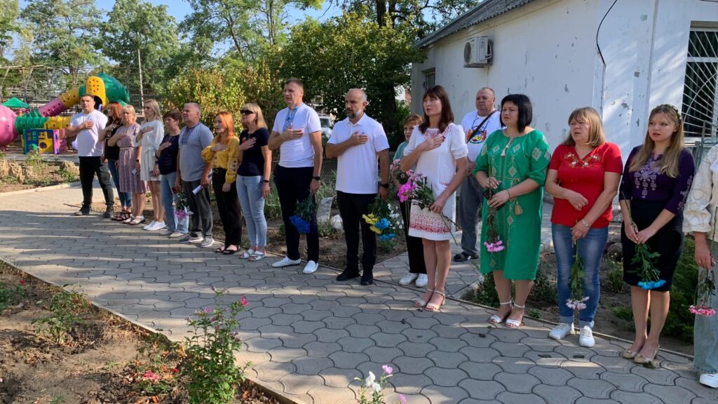 Група людей стоїть на вулиці, кожен тримає квіти й приклав руку до грудей. Позаду — дитячий майданчик із яскравими гірками та зеленими насадженнями. Атмосфера урочиста, схожа на вшанування пам’яті або громадську церемонію.