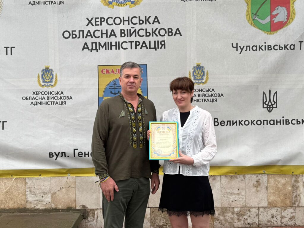 Михайло Линецький та ще одна особа стоять перед банером Херсонської обласної військової адміністрації. Вони тримають сертифікат із гербом України. На банері — назви населених пунктів, герб Скадовська та офіційна символіка.