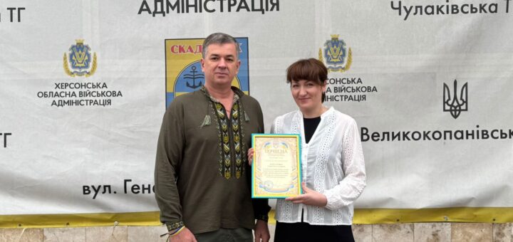 Михайло Линецький та ще одна особа стоять перед банером Херсонської обласної військової адміністрації. Вони тримають сертифікат із гербом України. На банері — назви населених пунктів, герб Скадовська та офіційна символіка.