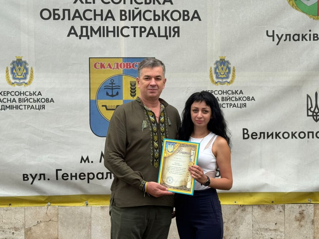 Михайло Линецький та ще одна особа стоять перед банером Херсонської обласної військової адміністрації. Вони тримають сертифікат із гербом України. На банері — назви населених пунктів, герб Скадовська та офіційна символіка.