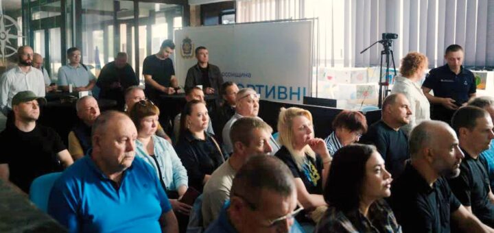 Група людей сидить у кімнаті, уважно слухаючи презентацію або доповідь. На передньому плані — камера на штативі, що фіксує подію. Позаду — банер із частково видимим написом «асоціація АКТИВН». У кімнаті — великі вікна з вертикальними жалюзі, світлі стіни та формальна атмосфера.