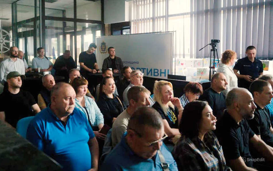 Група людей сидить у кімнаті, уважно слухаючи презентацію або доповідь. На передньому плані — камера на штативі, що фіксує подію. Позаду — банер із частково видимим написом «асоціація АКТИВН». У кімнаті — великі вікна з вертикальними жалюзі, світлі стіни та формальна атмосфера.