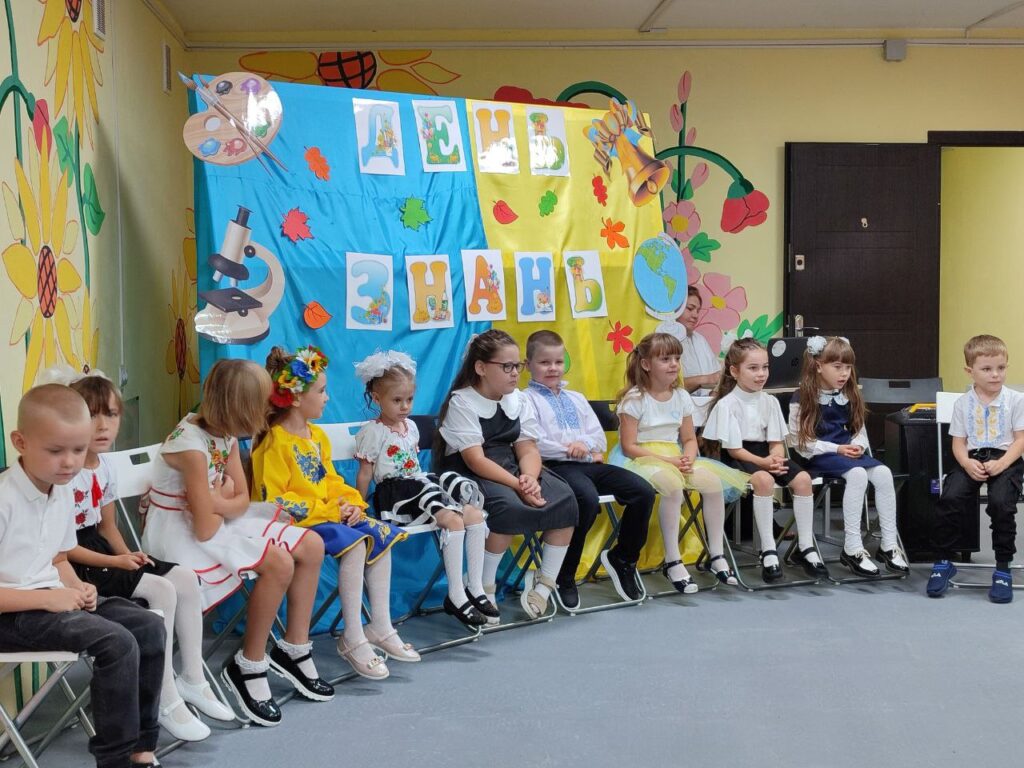 Група дітей у святковому та традиційному українському вбранні сидить у ряд на стільцях у класі. Позаду — яскраво оформлена стіна з написом «ДЕНЬ ЗНАНЬ», квітами, глобусом і мікроскопом. Атмосфера урочиста, початок навчального року.