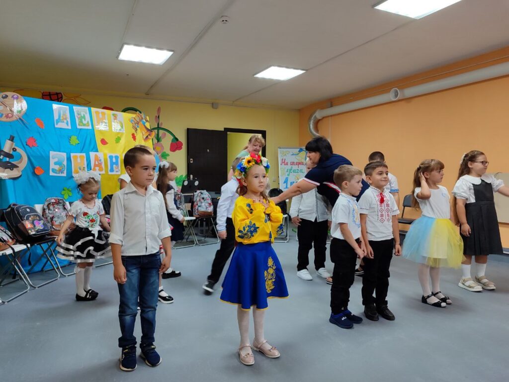 Група дітей у святковому та традиційному українському вбранні стоїть у класі перед стіною з написом «ДЕНЬ ЗНАНЬ». Дівчинка на передньому плані — у жовтій вишитій сорочці, синій спідниці з орнаментом і квітковому віночку. Позаду — повітряні кульки, паперові прикраси та шкільні рюкзаки на стільцях. Атмосфера урочиста, початок навчального року.