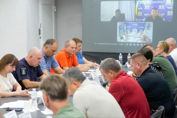 Група людей сидить за конференційним столом, беручи участь у гібридній зустрічі. На столі — документи, пляшки з водою, блокноти та іменні таблички. На великому екрані — два відеовікна з учасниками, які приєдналися дистанційно. Дехто вдягнений у форму або офіційний одяг. Атмосфера — ділова, організована.