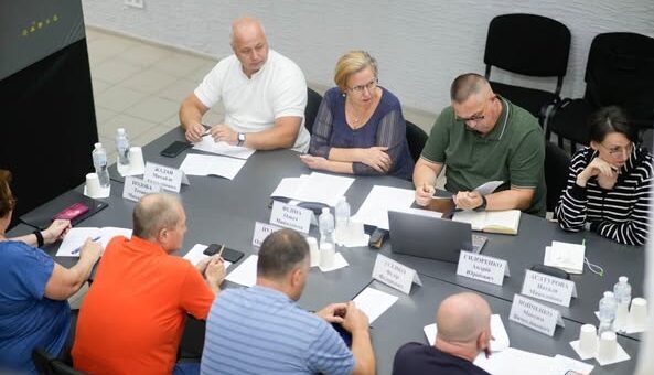 Група людей сидить за прямокутним столом у конференц-залі. Перед кожним — іменна табличка, документи, ноутбуки та пляшки з водою. Дехто користується телефонами. На задньому плані — світлі стіни, порожні стільці та плитка на підлозі. Атмосфера — офіційна, робоча.