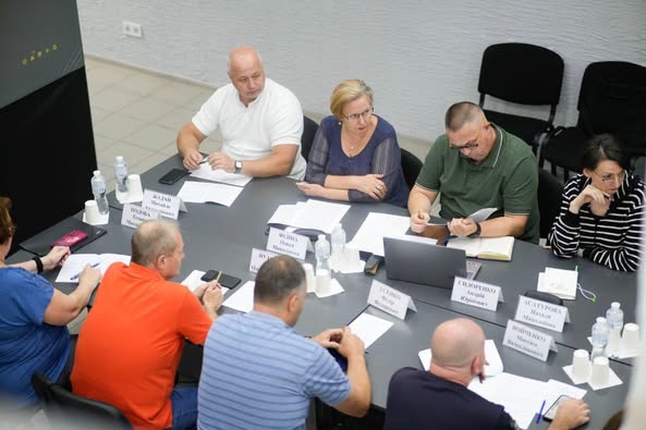 Група людей сидить за прямокутним столом у конференц-залі. Перед кожним — іменна табличка, документи, ноутбуки та пляшки з водою. Дехто користується телефонами. На задньому плані — світлі стіни, порожні стільці та плитка на підлозі. Атмосфера — офіційна, робоча.