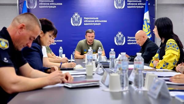 Група людей бере участь у нараді в кімнаті з синім банером «Херсонська обласна військова адміністрація». За столом — учасники в формі, цивільному одязі та традиційному українському вбранні. На столі — документи, пляшки з водою, іменні таблички. Олександр Прокудін веде обговорення.