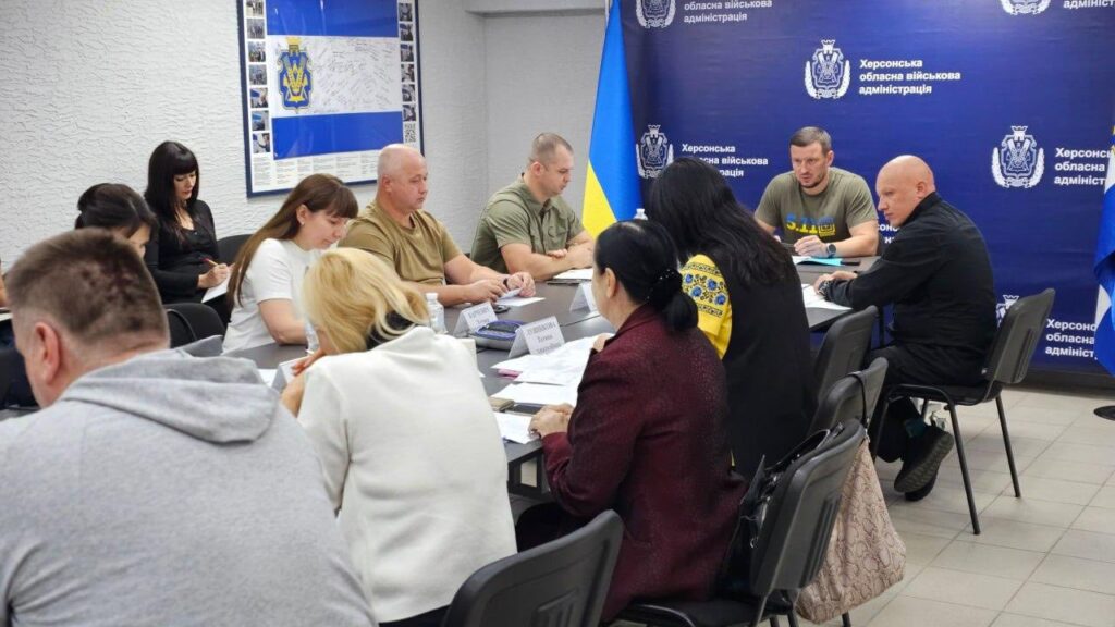 Група людей бере участь у нараді в кімнаті з синім банером «Херсонська обласна військова адміністрація». За столом — учасники в формі, цивільному одязі та традиційному українському вбранні. На столі — документи, пляшки з водою, іменні таблички. Олександр Прокудін веде обговорення.