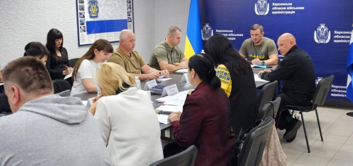 Група людей бере участь у нараді в кімнаті з синім банером «Херсонська обласна військова адміністрація». За столом — учасники в формі, цивільному одязі та традиційному українському вбранні. На столі — документи, пляшки з водою, іменні таблички. Олександр Прокудін веде обговорення.