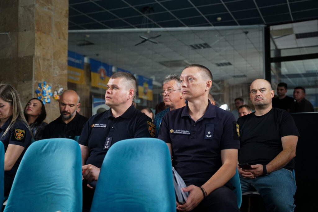 Група людей сидить у кімнаті з синіми кріслами, слухаючи презентацію або доповідь. Декілька осіб у передньому плані вдягнені у темну форму з емблемами, ймовірно представники служби безпеки або надзвичайних служб. Позаду — скляна стіна та плакати в синьо-жовтих кольорах. 