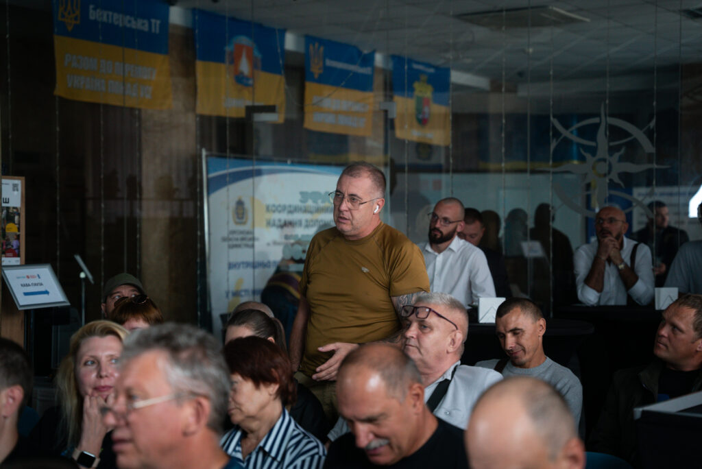 Група людей сидить у кімнаті, слухаючи виступ або презентацію. Один чоловік у зеленій сорочці стоїть, звертаючись до присутніх. На скляній стіні позаду — банери з українською символікою та написами: «Бахмутська ТГ», «Разом до перемоги України і над ворогом», «Координаційний штаб з питань соціального забезпечення військовослужбовців, військовозобов’язаних та членів їх сімей». Атмосфера — офіційна, патріотична.