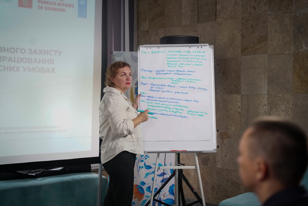 Жінка стоїть біля дошки з маркером, проводить презентацію в кімнаті з кам’яними стінами. На дошці — текст українською про цінності сім’ї, написаний синім і зеленим маркером. Ліворуч — екран із логотипами МЗС Данії та ПРООН. У передньому плані — чоловік, який слухає.