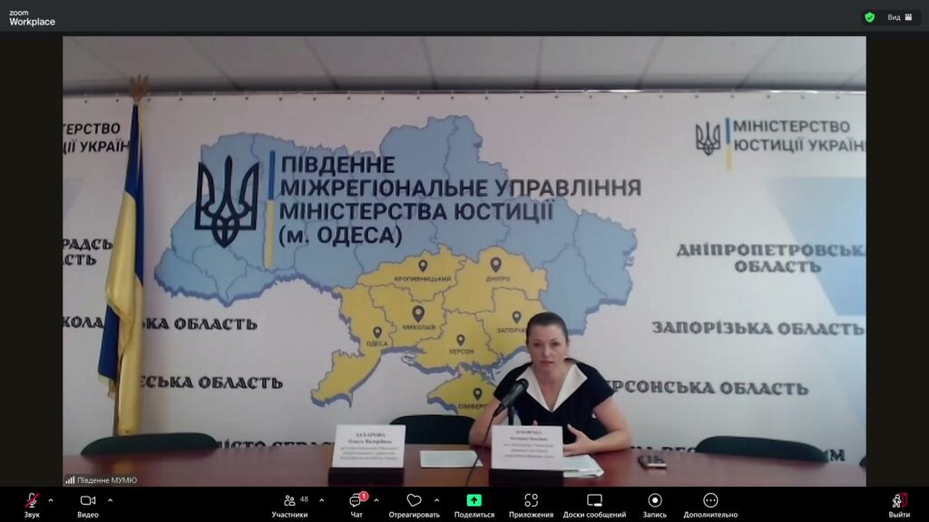 Жінка бере участь у Zoom-зустрічі, сидить за столом перед великою картою південних областей України. На стіні — емблема Міністерства юстиції України та напис: «ПІВДЕННЕ МІЖРЕГІОНАЛЬНЕ УПРАВЛІННЯ МІНІСТЕРСТВА ЮСТИЦІЇ (М. ОДЕСА)».