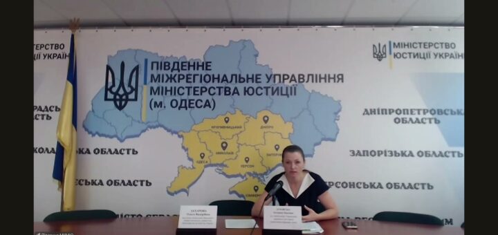 Жінка бере участь у Zoom-зустрічі, сидить за столом перед великою картою південних областей України. На стіні — емблема Міністерства юстиції України та напис: «ПІВДЕННЕ МІЖРЕГІОНАЛЬНЕ УПРАВЛІННЯ МІНІСТЕРСТВА ЮСТИЦІЇ (М. ОДЕСА)».