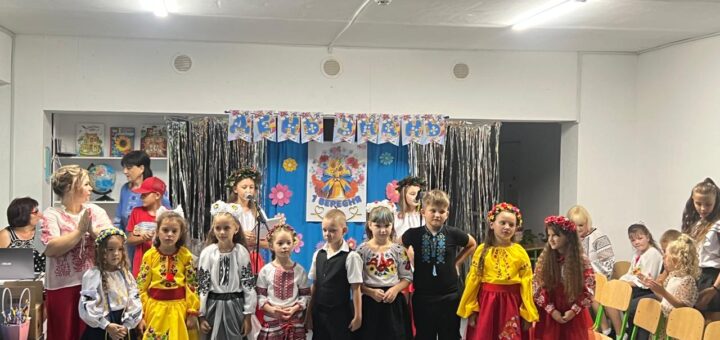 Група дітей у традиційному українському вбранні стоїть у ряд перед яскраво оформленою стіною з написом «ДЕНЬ ЗНАНЬ». Дівчатка — у вишиванках, квіткових віночках і кольорових спідницях. Позаду — дорослі, які спостерігають. Атмосфера святкова, схожа на шкільне свято.