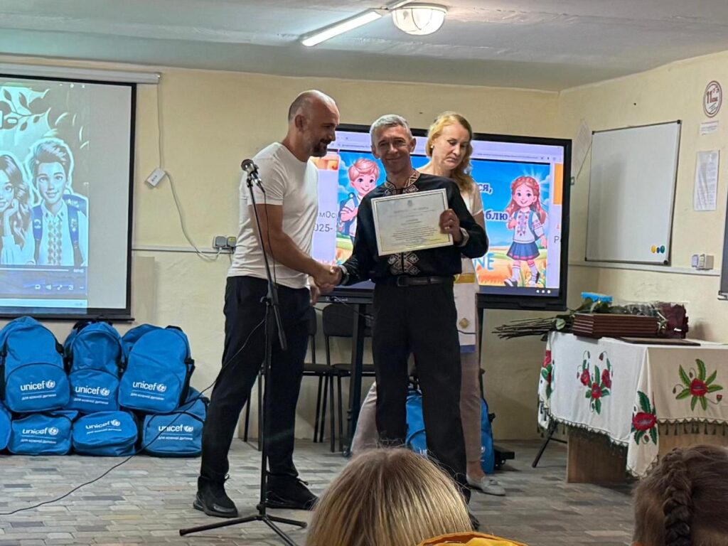 Троє дорослих беруть участь в церемонії вручення сертифікатів. Один чоловік передає документ іншому, який усміхається й демонструє його. Поруч — жінка, яка спостерігає. Позаду — два екрани з яскравими дитячими ілюстраціями та написами українською. Ліворуч — рюкзаки з логотипом ЮНІСЕФ, праворуч — стіл із вишитою скатертиною та згортками документів.