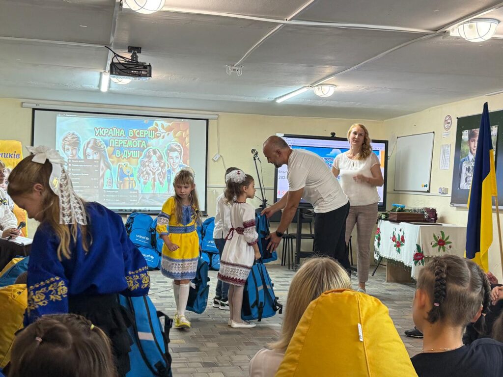 Чоловік передає рюкзаки двом дівчаткам у традиційному українському вбранні. Поруч — жінка, яка допомагає. У кімнаті — дорослі й діти, частина сидить, частина стоїть. На фоні — екран із написом «Україна в серці! Перемога в душі!» та зображенням дітей і прапора. Декор — українські прапори й вишиті рушники.