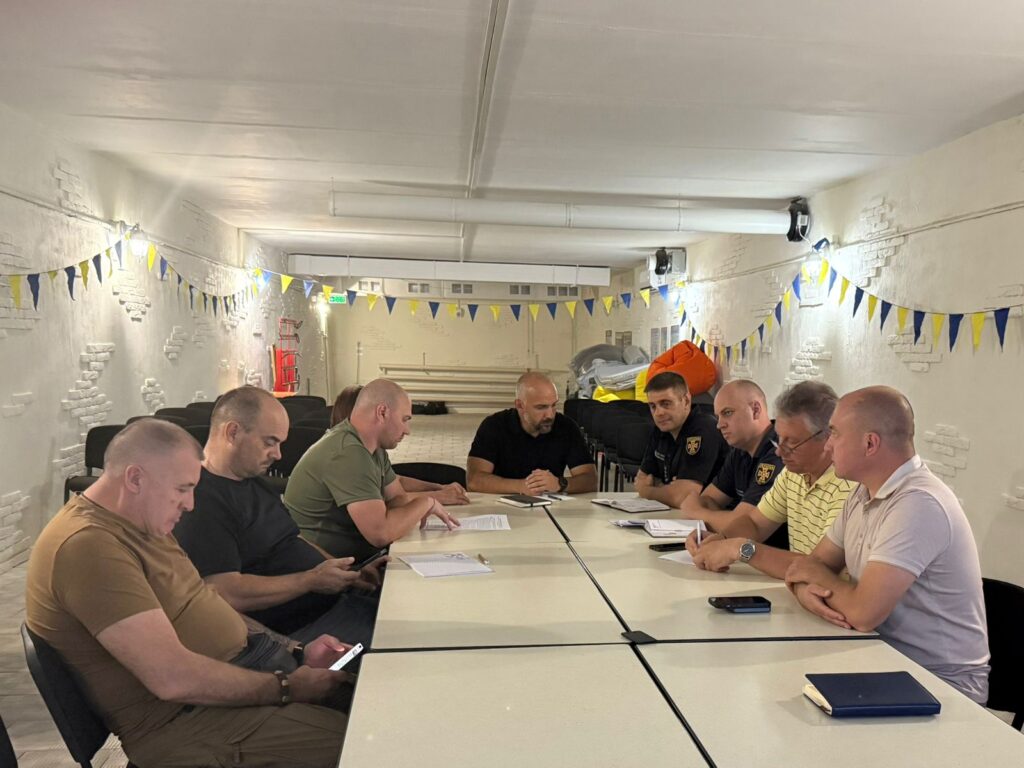 Дев’ятеро дорослих сидять за білими столами, розташованими прямокутником, у кімнаті з білими цегляними стінами. Декор — синьо-жовті прапорці, що натякають на українську тематику. Люди вдягнені у повсякденний та напівофіційний одяг, працюють із документами та електронними пристроями. 