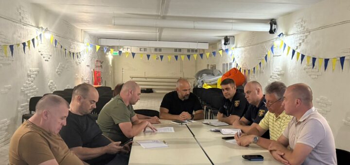 Дев’ятеро дорослих сидять за білими столами, розташованими прямокутником, у кімнаті з білими цегляними стінами. Декор — синьо-жовті прапорці, що натякають на українську тематику. Люди вдягнені у повсякденний та напівофіційний одяг, працюють із документами та електронними пристроями.