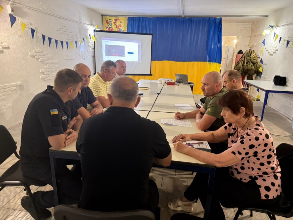 Дев’ятеро дорослих сидять за прямокутним столом у кімнаті з українськими прапорами та гірляндами. На екрані — слайд із діаграмою, що свідчить про презентацію. На столі — документи, ручки, блокноти. У фоні — військова форма на вішалці, біла стіна з текстурою та плитка на підлозі. 