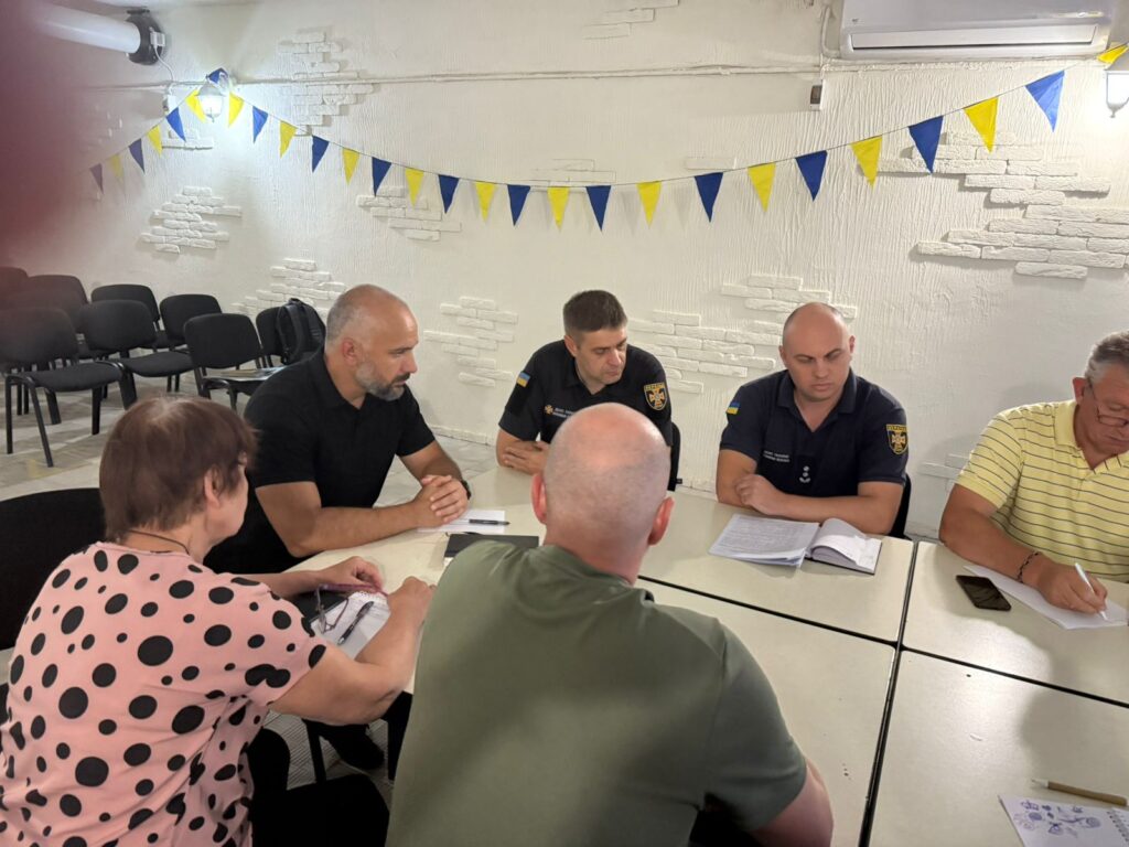 Сім дорослих сидять за білими столами, розташованими квадратом, обговорюючи документи. Двоє — у темній формі з емблемами, решта — в цивільному одязі. На стіні — біло-жовто-сині прапорці, що створюють урочисту атмосферу. Позаду — порожні чорні стільці, біла цегляна стіна та плакати.