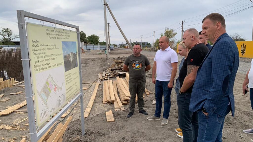 Шестеро дорослих стоять біля інформаційного щита на будівельному майданчику. Щит містить текст українською про проєкт будівництва дитячого садка на 120 місць у селі Новоолександрівка, Дніпровського району. На задньому плані — земля, дерев’яні дошки, будівельні матеріали, електроопори та будівлі. Атмосфера — робоча, інспекційна.