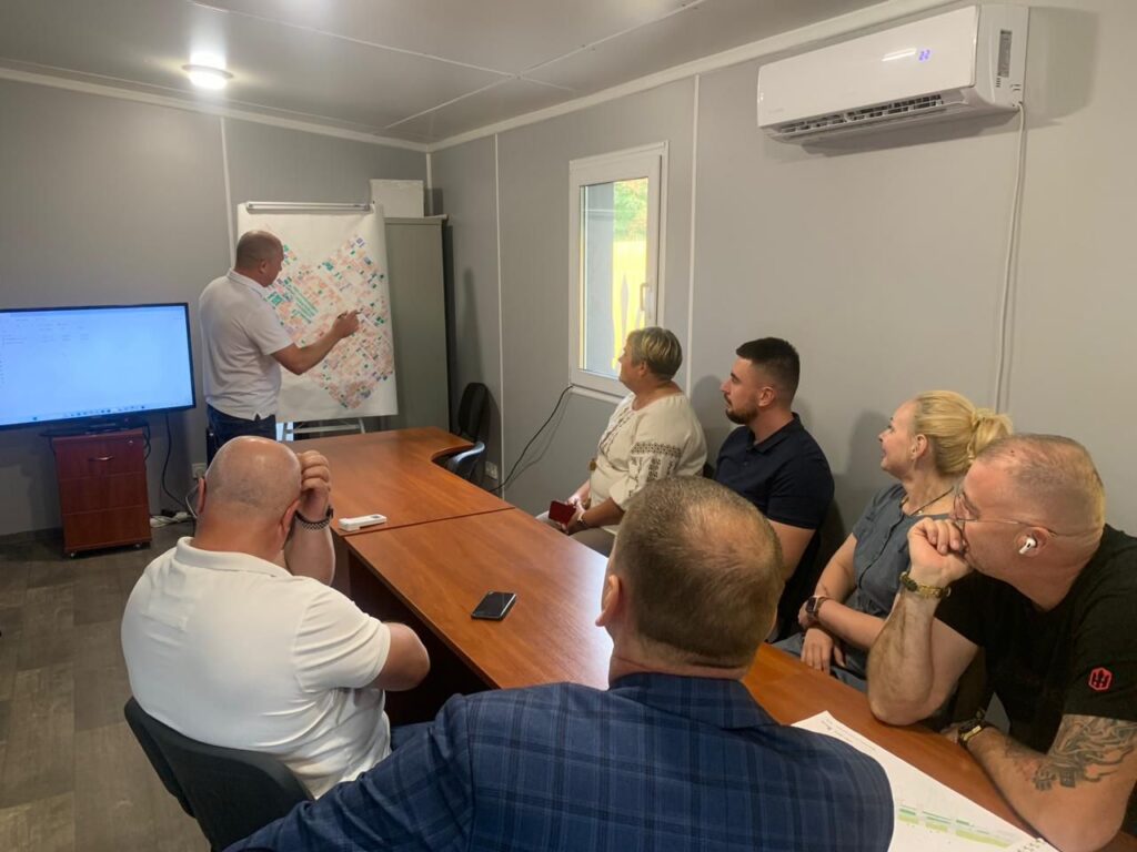 Група людей бере участь у зустрічі в кімнаті з сірими стінами. Один учасник стоїть біля фліпчарту з діаграмою або картою, пояснюючи зміст. На столі — документи, ноутбуки та матеріали з подібними схемами. Позаду — великий екран для презентацій. Атмосфера — робоча, стратегічна.