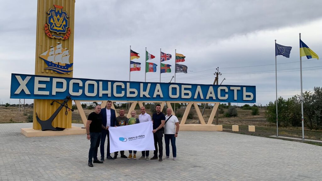Сім людей стоять перед великим знаком «ХЕРСОНСЬКА ОБЛАСТЬ». Позаду — жовта стела з гербом і стилізованим кораблем, а також прапори України, ЄС та інших організацій. Люди тримають білий банер із логотипом і текстом, що частково читається. Атмосфера — урочиста, можливо, пов’язана з візитом або громадською ініціативою.