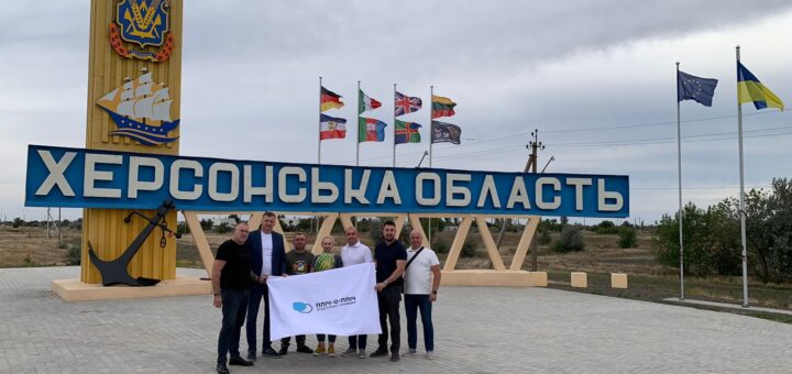 Сім людей стоять перед великим знаком «ХЕРСОНСЬКА ОБЛАСТЬ». Позаду — жовта стела з гербом і стилізованим кораблем, а також прапори України, ЄС та інших організацій. Люди тримають білий банер із логотипом і текстом, що частково читається. Атмосфера — урочиста, можливо, пов’язана з візитом або громадською ініціативою.