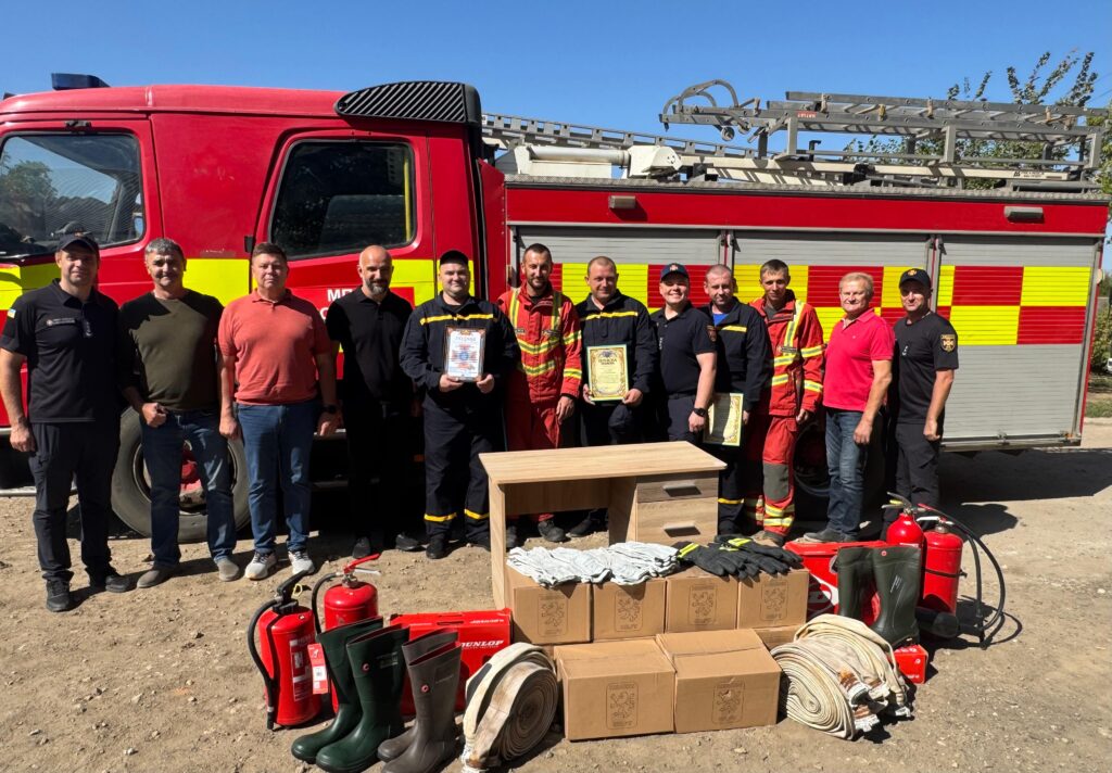 Тринадцять людей стоять перед червоною пожежною машиною з жовто-червоними смугами та драбиною. Більшість — у формі, дехто — у цивільному одязі. Двоє тримають сертифікати. Перед групою — вогнегасники, рукави, гумові чоботи, рукавички та картонні коробки. Атмосфера — урочиста, пов’язана з нагородженням або передачею спорядження.